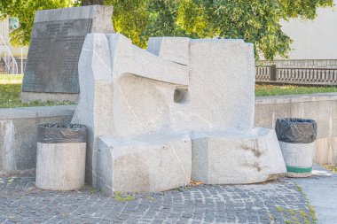 Yeşil bir ağaç ve binanın yanındaki kaldırım taşı zemininde soyut taş heykel, yakınındaki çöp tenekesi üç boyutlu doku ve zıt renkler