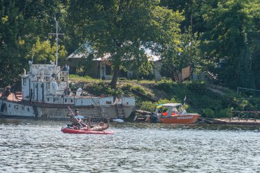 Rıhtım bölgesinde paslı vinci olan büyük bir mavna. Daha küçük turuncu motorlu tekne. Sakin su yansıması. Bitki örtüsünün doğal sınırı.