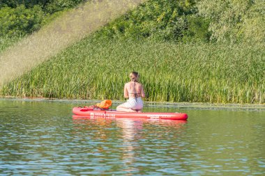 Sakin suda kırmızı kano, oturanlar, açık renk şort, turuncu can yeleği, yeşil kamış, güneşli gün, uzun gölgeler, doğal ortam Kyiv, Ukrayna 07-12-2025