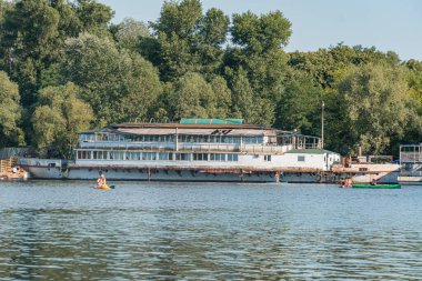 Kyiv rıhtımında çok katlı bir feribot, sessiz yeşil bitki örtüsü ve gri yapılar ile, yumuşak sabah veya geç saatlerde Ukrayna, Kyiv, 07-12-2025 ışıklandırması altında