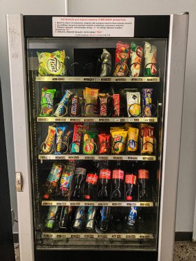 Ukrayna 'da iyi stoklanmış bir yiyecek otomatının önünde duran kişi cam kapıya yansıyor. Cipsler, gazozlar, paketlenmiş eşyalar birden fazla rafta. Bright i Kyiv, Ukrayna 07-08-2025