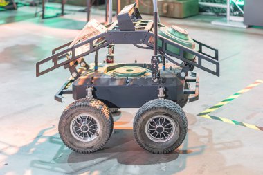 Görüntü dört tekerli gri bir yer dronu gösteriyor. Çekiş tekerleri ve geniş manipülatör kolu var. Endüstriyel ortamda çalışıyor.