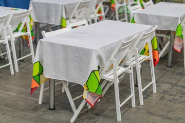 Geniş masa oyun odası, renkli tabaklar, beyaz masalar, sandalyeler, soyut duvar sanatı ve beton zemin, alanı aydınlatan doğal ışık.