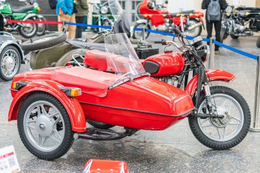 Araba fuarında sepetli klasik tasarımı ve krom detaylarıyla iyi korunan klasik kırmızı motosiklet. Arka planda çeşitli renkli araçlar var. Beyaz çizgili mavi bir araba.