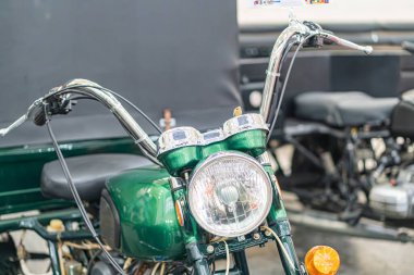 Klasik yeşil motosiklet, loş ışıklandırma, ön üç çeyrek manzara, krom aksanlar, detaylı tasarım, desenli ve eğimli yüzeyler, parlak yansımalar, işçilik odaklı, promosyon veya ed