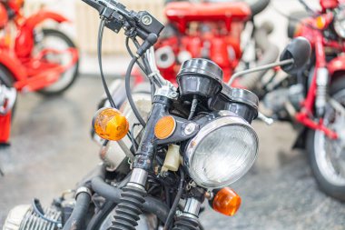 Kapalı mekanda park etmiş, klasik kırmızı bir motosikletin yakından görüntüsü. Motosikletin metalik bir far çerçevesi, gidon kontrolleri ve ızgara desenli motosikletleri var.