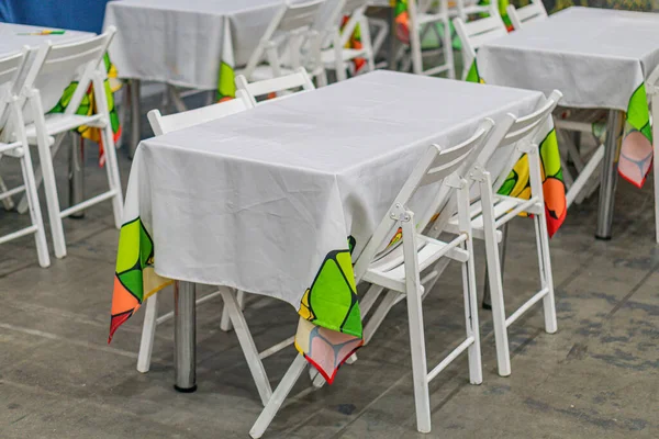 Geniş masa oyun odası, renkli tabaklar, beyaz masalar, sandalyeler, soyut duvar sanatı ve beton zemin, alanı aydınlatan doğal ışık.