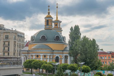 Mavi çatılı canlı sarı-yeşil bir kilise Ukrayna 'nın başkenti Kyiv' in arka planına karşı ayakta duruyor. Birden fazla kubbesi var, biri altın haçla kaplı, ve ben Kyiv, Ukrayna 07-06-2025