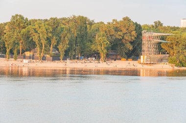 Sakin Kyiv plaj ahşap rıhtımı, dağınık tekneler, inşaat alanı, bereketli ağaçlar, turuncu ve mavi renkler, uzun gölgeler, gerçekçi stil Kyiv, Ukrayna 07-06-2025