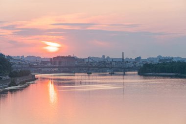 Nehir üzerinde gün batımı, Kyiv silueti, birden fazla kemerli köprü, sakin su gövdesi, şehir manzarası, turuncu ve pembe tonlar, serin maviler ve yeşiller, düşük aydınlatma Kyiv, Ukrayna 07-06-2025