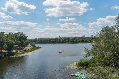 Partly cloudy sky over calm lake in Kyiv, Ukraine Boats red canoe, greenvegetation covered boat on water Lush greenery at shoreline Kyiv, Ukraine 07-12-2025