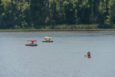 Hareket eden kanolar, yumuşak ışıklandırma, durgun su yansıtan yeşil ve pastel gökyüzü, kırmızı ve sarı bayraklar, uzak durgun tekne, Kyiv, Ukrayna 07-12-2025