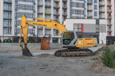 Bir inşaat alanında çalışan büyük sarı kazıcı, yakınlardaki toprak yığını, arka planda bitmemiş beyaz ve siyah binalar, kırmızı kamyon, bulutlu gökyüzü Kyiv, Ukrayna 07-12-2025
