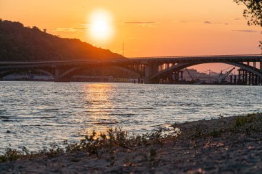 Ukrayna, Kyiv 'de bir nehir tarafından gün batımı Sıcak renkleri yansıtan sakin su, birden fazla kemerli köprü, ön planda kayalar, hafif aydınlatma Kyiv, Ukrayna 05-31-2025