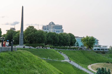 Gündoğumunda Kyiv parkında bulunan dikilitaş, yeşil çimenler, ağaçlar, beyaz adımlar, çağdaş bina, sıcak aydınlatma, yumuşak renkler, nazik sis, dengeli bileşim Kyiv, Ukrayna 06-01-2025