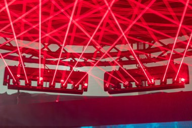 Kırmızı ışık ve spot ışıklarının hakim olduğu karanlık bir sahnenin düşük açılı çekimi endüstriyel bir DJ kabininin üzerinde desenler yaratıyor.