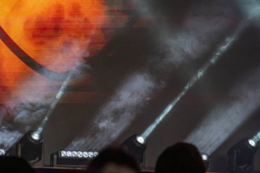 Canlı bir konser sahnesinin geniş görünümü, sıcak kehribar sahne ışıklandırması ile serin mavi arkaplan ile karşıt merkez ekran dinamik soyut grafikler görüntüler, seyirci odaklı sahne ışıkları Ove tarafından çevrili