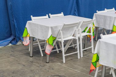 Düzenlenmiş yemek masaları, beyaz masa örtüleri, renkli masa örtüleri, uyumlu sandalyeler ve oditoryum ya da etkinlik alanını gösteren mavi kumaşla kaplı bir duvar olan kapalı masa oyunu odası.