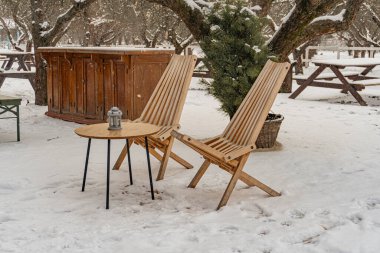 Kış sahnesi, karla kaplı zemin, verandada iki açılı Adirondack sandalyesi, mumlu küçük yuvarlak masa, sabah ya da akşam aydınlatması, dış mobilya ve yapılar, bir dizi