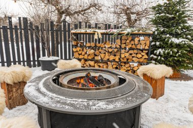 Koyun derisi yastıklarla çevrili modern kış ateşi çukuru ve arka plandaki karlı ağaçların içinde parlayan kırsal çitlerle çevrili.