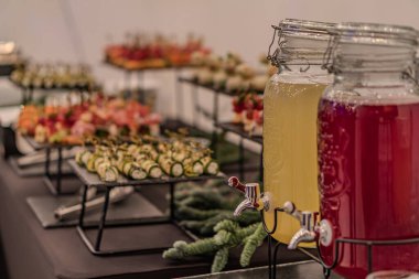 Rustic table with a white cloth, holding two sealed glass jars one pinkish-purple liquid, one yellowish, pickled vegetables in wire racks various colors green, orange, red, soft shadows, warm to