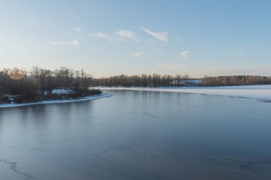 Sakin kış gölü, kıyı şeridinde donmuş buz, yumuşak açık mavi, gri, uzun gölgeler, muhtemel şafak veya alacakaranlık, bulutsuz açık gökyüzü, yapraksız uzak ağaçlar, seyrek bitki örtüsü, dalgalar, kırık buz p