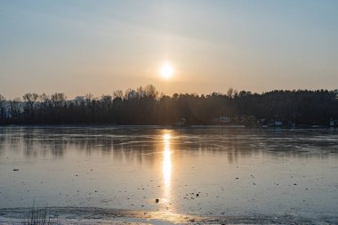 Buz ve karla kısmen kaplanmış sakin bir göl sahnesi. Alçak güneş, çorak gökyüzünün altında ılık bir parıltı yayıyor. Titreşimden gelen buz renklerini yansıtıyor.