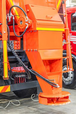 Altında büyük turuncu inşaat aracı, çoklu tekerlekler, merkezi dingil, mekanik bileşenler, hidrolik hatlar, endüstriyel ortam, kapalı atölye veya garaj, karmaşık tasarım, yapay ışık