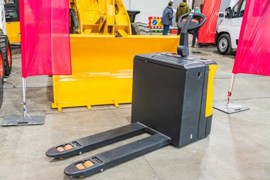 Sarı inşaat aracı yan tarafta, siyah hidrolik eklenti, beton zemin, sanayi deposu ayarları, yüksek tavanlar, büyük makineler, iki kişi koyu renk, açık renk elbise, sent 'ten uzaklık