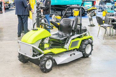 Kapalı alanda geniş yeşil engebeli bir çim biçme makinesine yakın çekim, kameraya dönük, arka planda konuşan iki kişi koyu renk ceketler ve pantolonlar giyerek iç mekanda