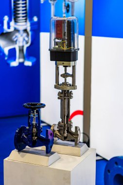 Beyaz kaidedeki merkezi metal yapı, bir sergi salonunun yapay ışığı altında parlayan sarmalanmış siyah bir hortum, çapraz gümüş borular ve dairesel cam muhafaza Ind.