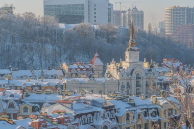 Kiev 'in kış manzarası, Ukrayna' nın karla kaplı çatı ve caddeleri, çeşitli mimari stiller, büyük kule, süslü dekorasyonlar, kalın kar tabakası, soluk renkler, çıplak ağaç dalları, bulutlu gökyüzü,