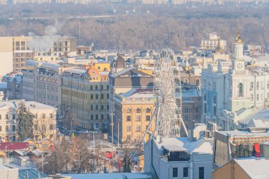 Bulutlu gökyüzü altındaki şehir manzarası, canlı kırmızı dönme dolap, klasik ve süslü düşük binalar, mavi altın aksanlı yapılar, karla kaplı sokaklar, demet halinde bireyler, yüksek vantilatörlü poi