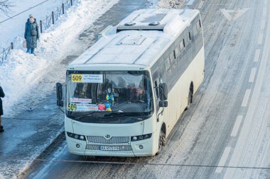 Ukrayna 'nın başkenti Kyiv' de karlı yolda ilerleyen şehir taşımacılık otobüsü. Büyük beyaz araç, içeriden görülebilen kullanım izleri gösteriyor. İki pedestri Kyiv, Ukrayna - 17 Şubat 2026