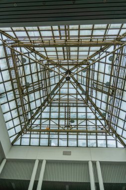 Çoklu kiriş ve kirişlerden oluşan merkezi geometrik kafes yapısına sahip geniş bir iç mekan, dikey direkler tarafından desteklenen yarı şeffaf tavan panelleri doğal ışığı uzaya filtreler Th
