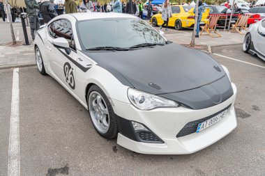 Siyah aksanlı, eşsiz bir vücut kiti, yarış şeritleri ve büyük tekerlekleri olan beyaz bir spor araba bir sokakta ya da otoparkta park edilmiş görünüyor. Olay yeri Gath Kyiv, Ukrayna 10-12-2024