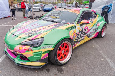 Bu resimde, canlı bir yarış arabası, muhtemelen bir yarış pistinde veya otomotiv etkinliğinde, HELIOS gibi sponsor logoları gösteriliyor ve Kyiv, Ukrayna 10-12-2024 için çok dilli tekerlekleri var.