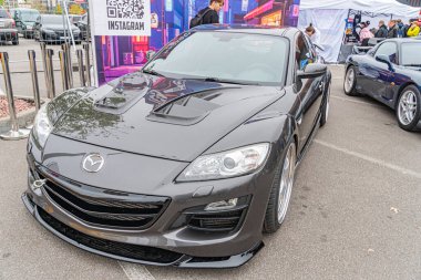 Siyah bir spor araba muhtemelen Mazda RX veya MX modeli, agresif tasarımı ve özelleştirilmiş alaşım tekerleklerini vurgulayan bir otomobil fuarında sergilenmektedir.
