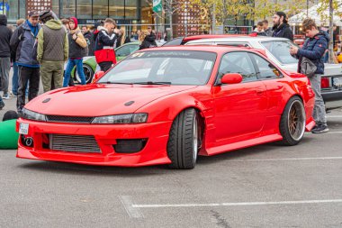 Geniş gövde kiti gibi modifikasyonları olan kırmızı bir spor araba, havalandırma kaputu, ve çok dilli satış sonrası tekerlekleri açık bir etkinliğe park edilmiş. Günlük kıyafetler giyen insanlar Kyiv, Ukrayna 10-12-2024