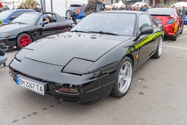 Tekerlekler, yan etekler ve arka kanat spoiler gibi özelleştirilmiş siyah bir spor araba, bir etkinlikte sergileniyor. Kenarlarında yeşil bir şerit var ve plakası Kyiv, Ukrayna 10-12-2024