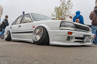 Kamuya açık bir etkinlikte muhtemelen sürüklenmek ya da yarışmak için kullanılan beyaz bir arabanın iki resmi park halindeki arabayı gösteriyor. İkinci fotoğraf ise Ukrayna, Kyiv, 10-12-2024 'ü gösteriyor.