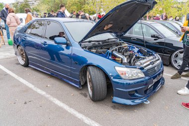 Arka pazar modifikasyonlarına sahip mavi bir sedan kaputu açık, muhtemelen açık bir alanda ağaçların yanında duruyor. Birkaç kişi etrafta dikiliyor, Ukrayna, Kyiv, 10-12-2024 ile meşgul.