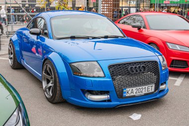 Meşgul bir şehir caddesinde mavi bir Audi spor arabası park edilmiş, büyük ızgara, LED farlar ve alaşımlı tekerlekler gibi belirgin özelliklerini sergiliyor.