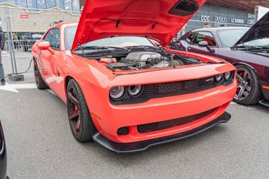 Açık hava ortamında, muhtemelen bir park alanında ya da sergi alanında siyah iç mekan ve büyük tekerlekleri olan parlak kırmızı bir Amerikan kaslı arabası sergilenmektedir.