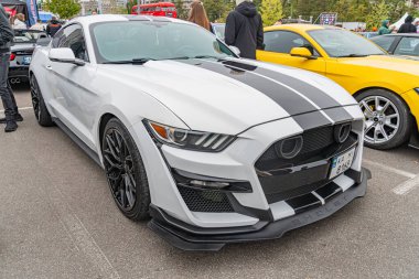Geniş gövdeli modifikasyonlara sahip beyaz ve siyah bir Ford Mustang, diğer arabaların arasında bir otoparkta sergileniyor.