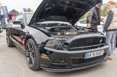 Açık kapüşonlu siyah bir Ford Mustang GT, muhtemelen bir araba yarışmasında, 50L V8 Ti-VCT motorunu sergiliyor. Kapalı alanda çekilmiş gizli bir fotoğraf ya da bulutlu bir günde Kyiv, Ukrayna, 10-12-2024