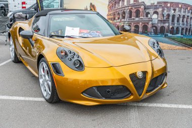 Görüntü, sarı bir Alfa Romeo Spider 'ın kaldırımlı bir sokakta ya da otoparkta park ettiğini gösteriyor. Alaşımlı tekerlekler ve aerodinamik tasarım içeriyor. Arka plan muhtemelen Kyiv, Ukrayna' dan 10-12-2024 model bulanık bir bina içeriyor.
