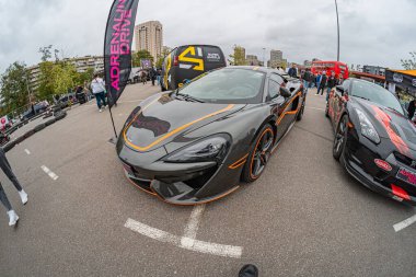 İki spor arabanın panoramik görüntüsü bir araba fuarında veya şehir ortamında sergileniyor. Siyah ve turuncu renkli spor araba çizgileri, büyük jantlar, aerodinamik tasarım Kyiv, Ukrayna 10-12-2024