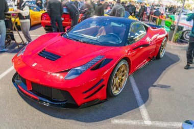 Bir otomobil fuarında ya da etkinlikte altın jantları ve parlak bir tasarımı olan canlı kırmızı bir spor araba bir grup insan ve diğer araçların arasında göze çarpıyor.