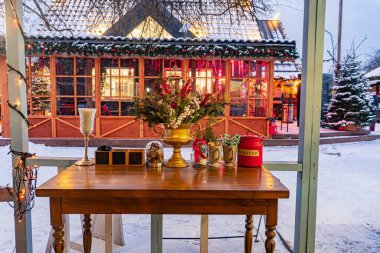 Kış ayarı, karla kaplı ön planda eski ahşap bir masa, kırmızı ve altın desenli kumaş, dekoratif eşyalar mumlar, altın beyazı, küçük ağaçlı re Kyiv, Ukrayna - 15 Şubat 2025